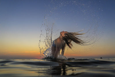 Girl splashing water with hair in sea against sky during sunset