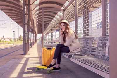 Full length of woman sitting with luggage on bench at railroad station platform