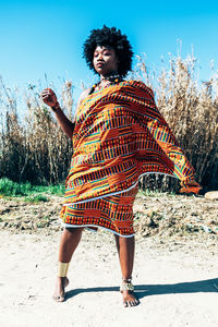 Serious african model with afro hairstyle wearing colorful handwoven kente and looking away while standing on field on bright summer day