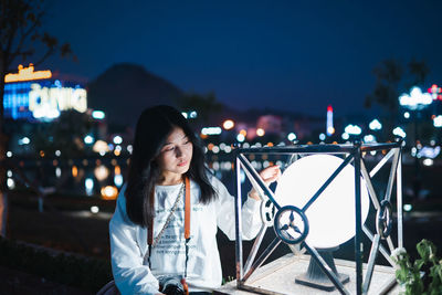 Young woman looking at illuminated light in city against sky