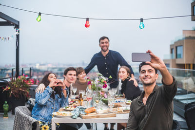 Smiling man taking selfie through smart phone while enjoying with friends in social gathering on building terrace