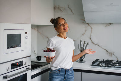 Pretty smiling young woman holds a plate of fresh cherry berries in her hand in the kitchen