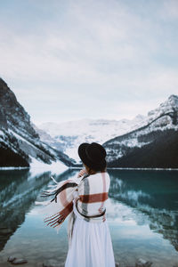 Back view of anonymous female in white dress and scarf standing towards clean water of lake louise against snowy mountain ridge on winter day in alberta, canada