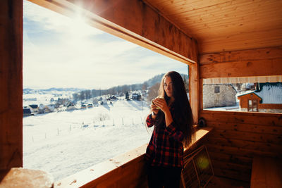 Portrait of young woman holding cup while standing by window in cottage during winter