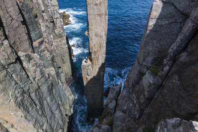 Woman climbs a free standing rock pilar which emerges from the ocean.