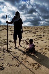 Full length of man holding stick while standing by girl on sand