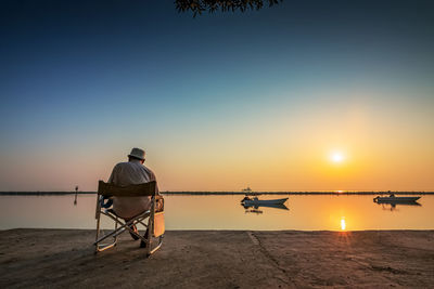 Rear view of man sitting on beach against sky during sunset
