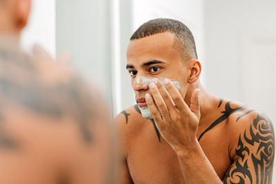 Tattooed shirtless man applying shaving cream on face while looking in mirror