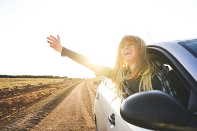 Young woman with arms outstretched leaning out from car window against clear sky