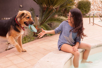 Teenage girl gesturing towards dog holding slipper in mouth at poolside