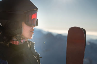 Portrait of sportswoman in helmet and mask with skis in hand, looking away, enjoying sunny frosty