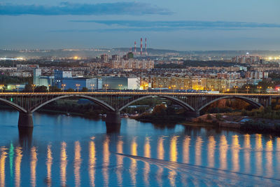 Illuminated bridge over river by buildings against sky in city