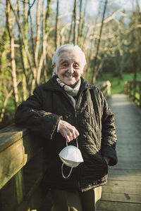 Smiling elderly woman on footbridge holding protective face mask during covid-19