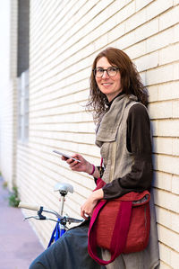 Smiling young woman with bike leaning on wall while using mobile