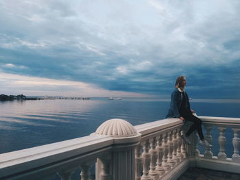 Woman sitting on railing against sea during sunset