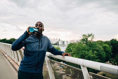 Portrait of thirsty sportsman drinking from bottle while standing on footbridge against sky