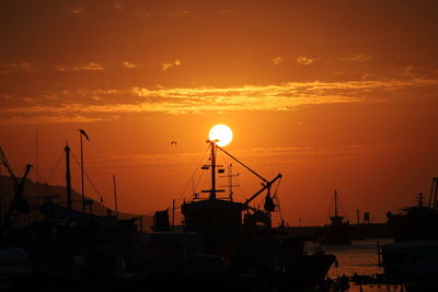 Silhouette cranes at harbor against orange sky