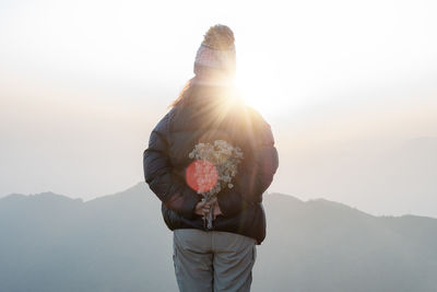 Rear view of woman holding flowers while standing against mountain and sky
