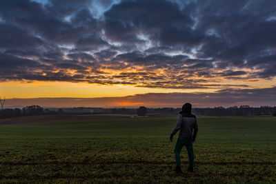 Rear view of man standing on field against cloudy sky during sunset