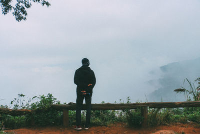 Rear view of man standing by railing and plants against sky