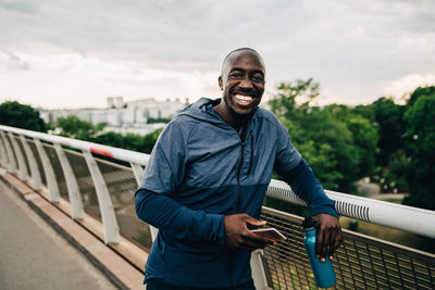 Portrait of cheerful sportsman holding mobile phone and bottle while standing on footbridge