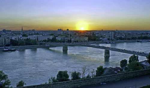 Bridge over river by buildings against sky during sunset