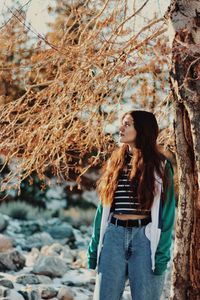 Young woman looking away while standing by bare tree during autumn