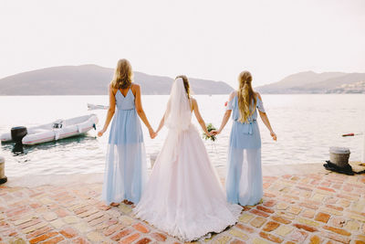 Rear view of bride holding hands with bridesmaids by sea against clear sky