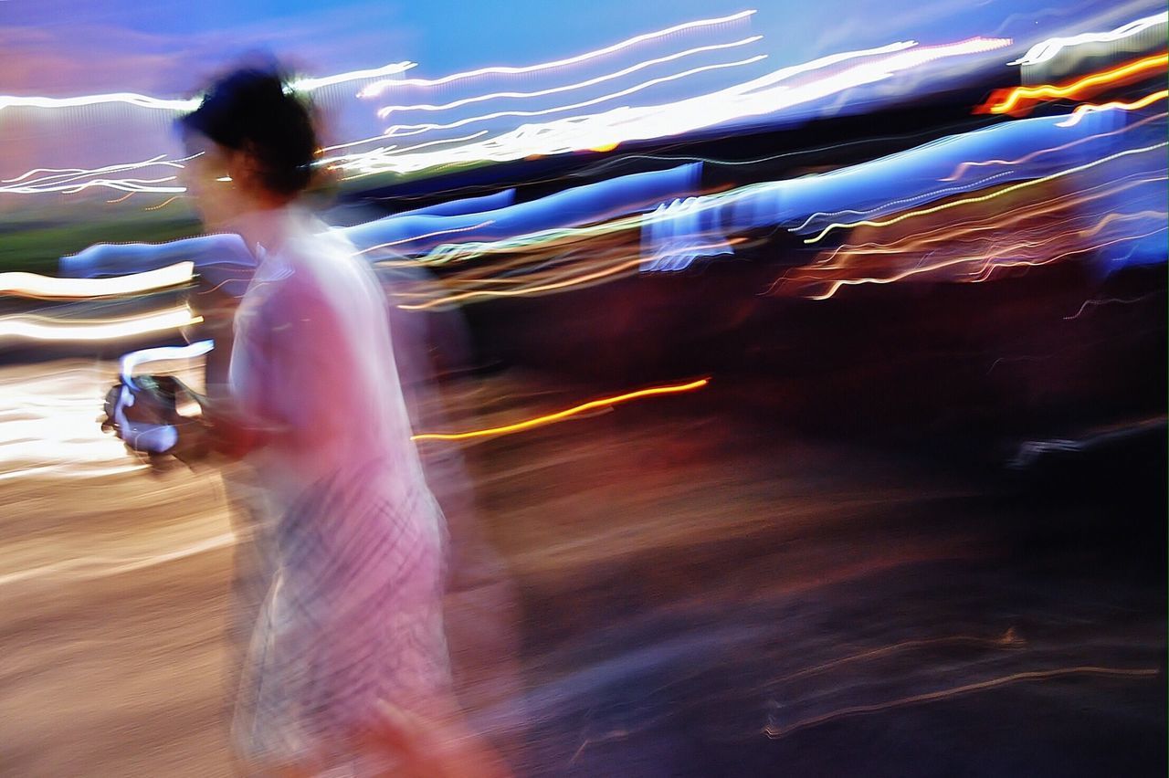 BLURRED MOTION OF WOMAN WALKING ON ILLUMINATED CITY AT NIGHT