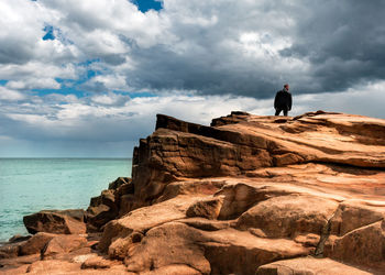 Man standing on rock by sea against cloudy sky