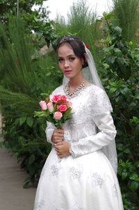 Portrait of beautiful bride holding flowers while standing against plants