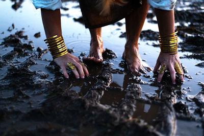 A woman's bare feet and hands with luxury bracelets and rings in a dirty puddle of black mud