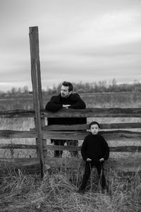 Father and son in black clothes stand in a field with an old wooden horse fence in the fall