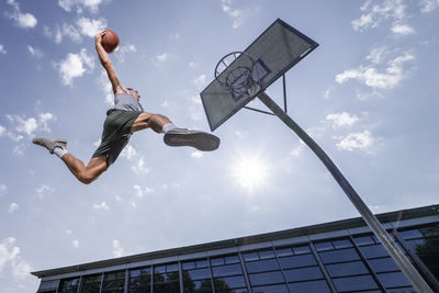 Young man dunking ball in hoop while playing basketball against sky on sunny day