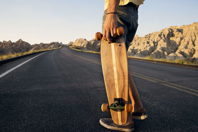 Low section of man holding skateboard while standing on country road on desert