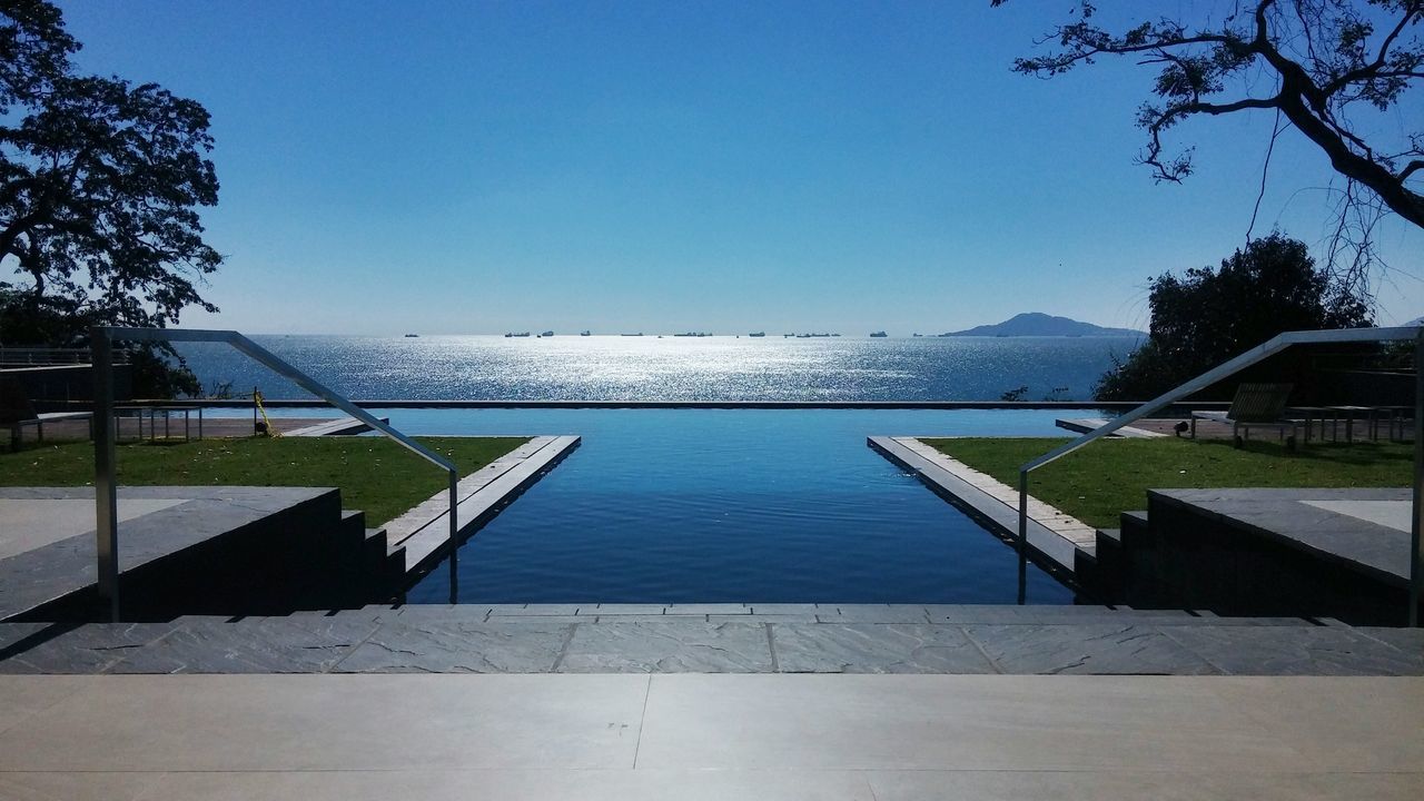 clear sky, blue, copy space, built structure, railing, tree, architecture, water, building exterior, tranquility, sunlight, nature, bench, tranquil scene, day, grass, outdoors, sky, empty, walkway