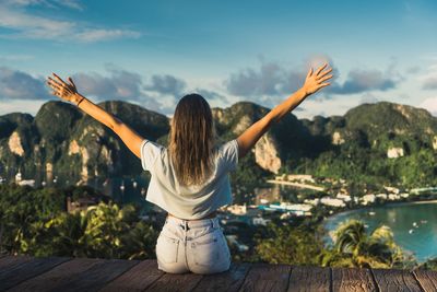 Woman with arms outstretched sitting on pier against sky