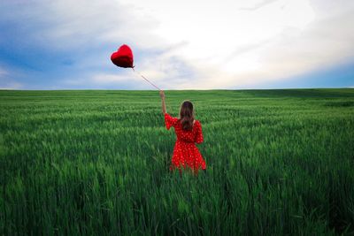 Woman in red dress standing in a field of green wheat and holding a red heart balloon