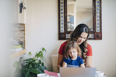 Daughter sitting while mother using laptop at home office