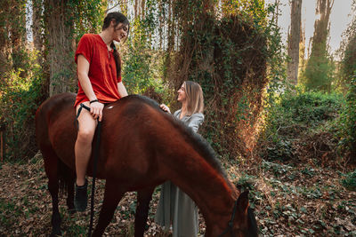 Young woman sitting on horse talking with friend in forest