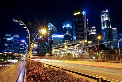 Light trails on road by illuminated buildings against sky at night