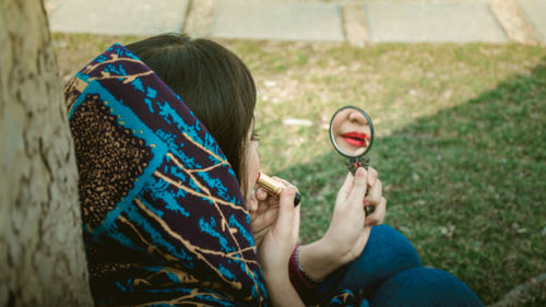 High angle view of woman applying lipstick while holding mirror on grass