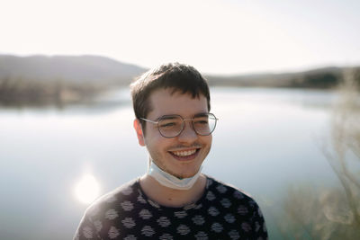 Young cheerful tourist in face mask and eyeglasses standing near river behind mountains while looking away