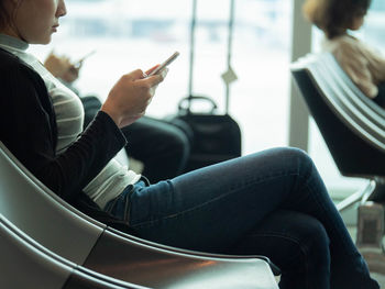Midsection of woman using mobile phone while sitting on chair at airport departure area