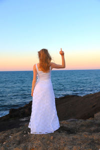 Rear view of woman showing middle finger while standing at beach against during sunset