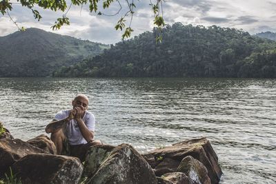 Mid adult man smoking while sitting on rock by lake against sky