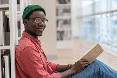 Black student man library visitor sitting on floor near bookcase with book in hands, looks at camera