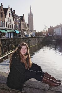 Portrait of smiling mid adult woman sitting on retaining wall over canal in city