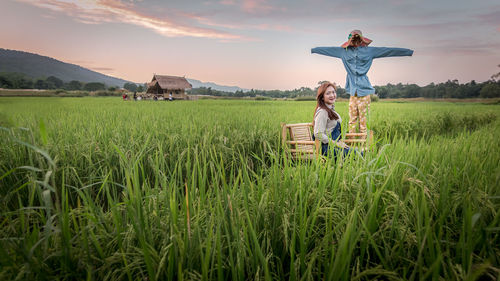 Portrait of woman sitting by scarecrow on grassy field against sky