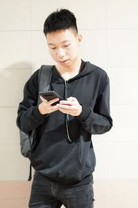 Young man using phone while standing against wall